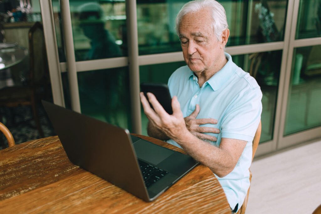 Elderly man using smartphone and laptop at wooden table indoors.