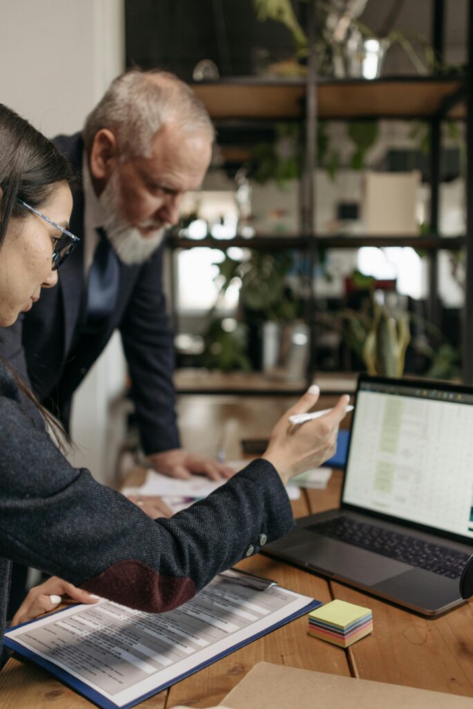 A business professional explains data to a colleague using a laptop and documents in an office setting.